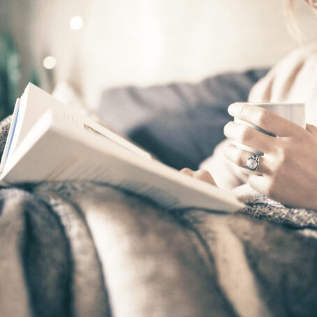 Image of a woman sitting on the couch, covered in a blanket, reading a book, and holding a mug of coffee.
