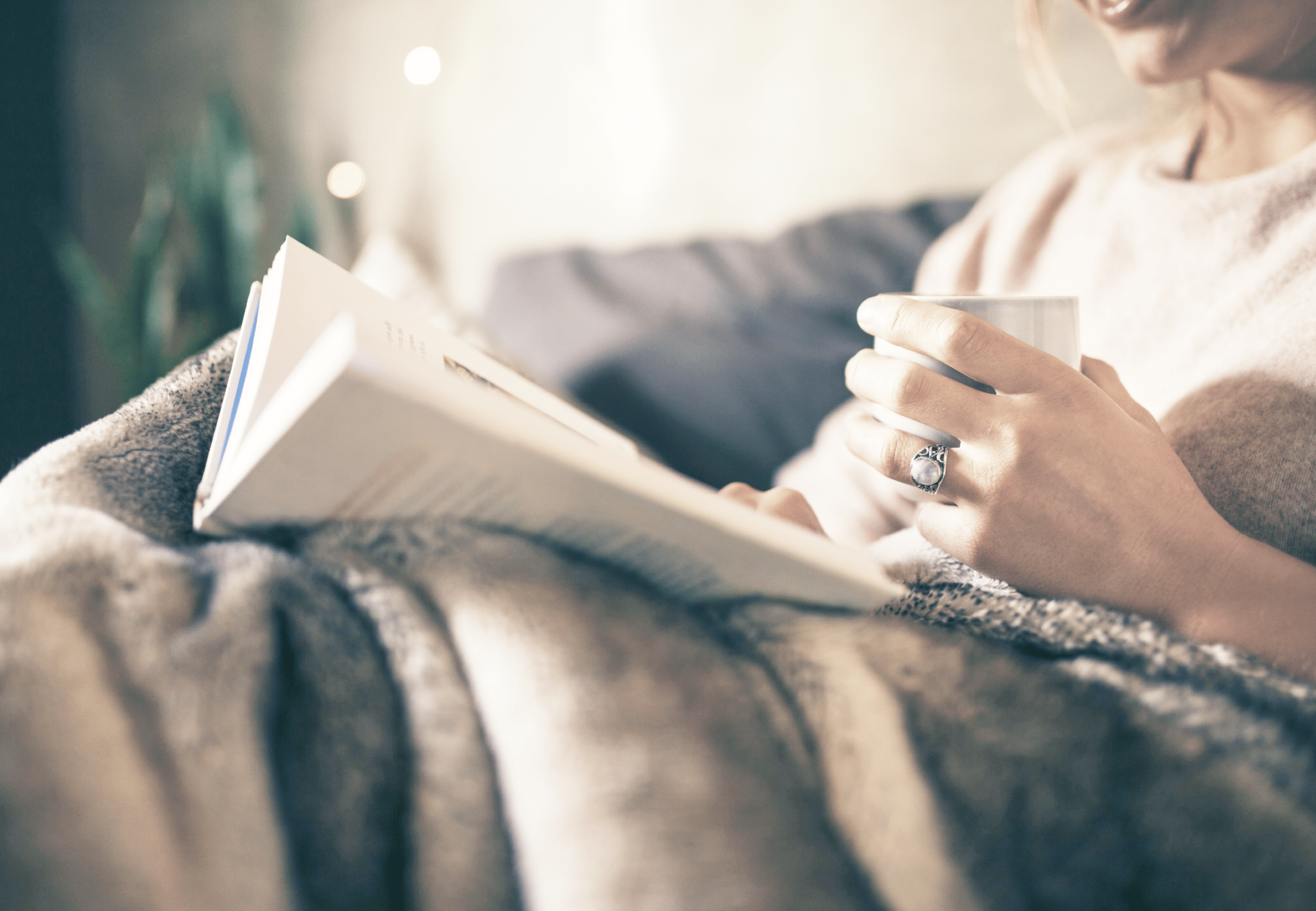 Image of a woman sitting on the couch, covered in a blanket, reading a book, and holding a mug of coffee.