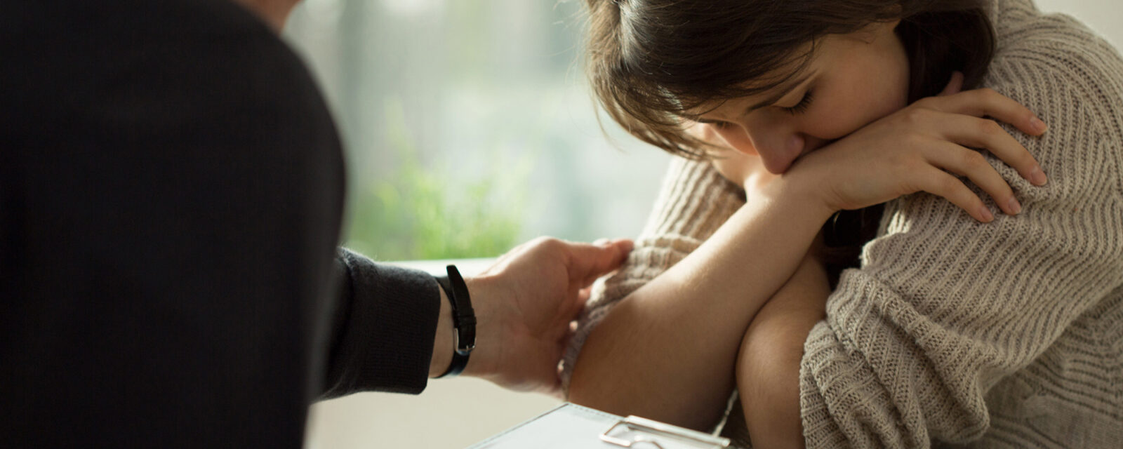 This image pictures a woman looking down with her arms crossed over her chest and being comforted by another person.