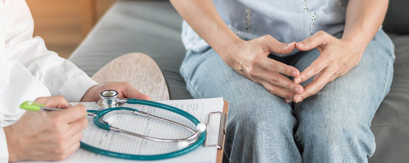Close up image of a doctor speaking with his patient. The doctor is holding a clip board with a stethoscope laying on top. The patient is holding her hands together.