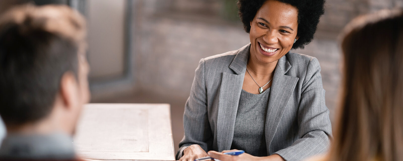 Image of a woman in a grey suit smiling at her two clients, who are out of focus in the image.