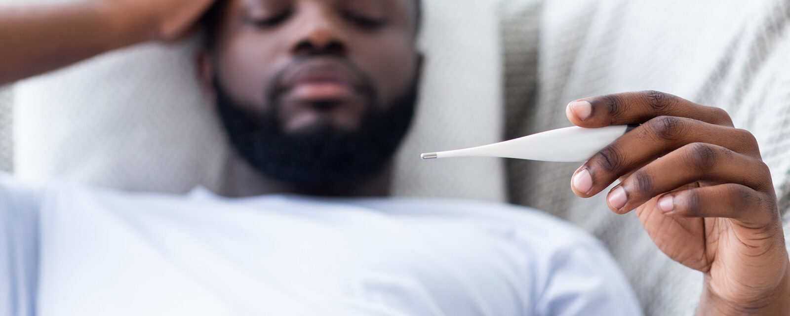 Image of a man laying in bed with his hand on his forehead and the other one holding a thermometer.