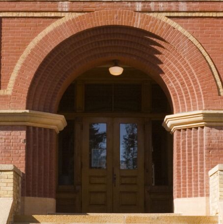 Horizontal straight-on exterior view of an old red brick high school or elementary schoolhouse building facade, centered on the entrance front door and staircase. The traditional architecture suggests a respect for higher education and learning. The brownstone built structure is located in a small Midwestern town in the U.S.A.