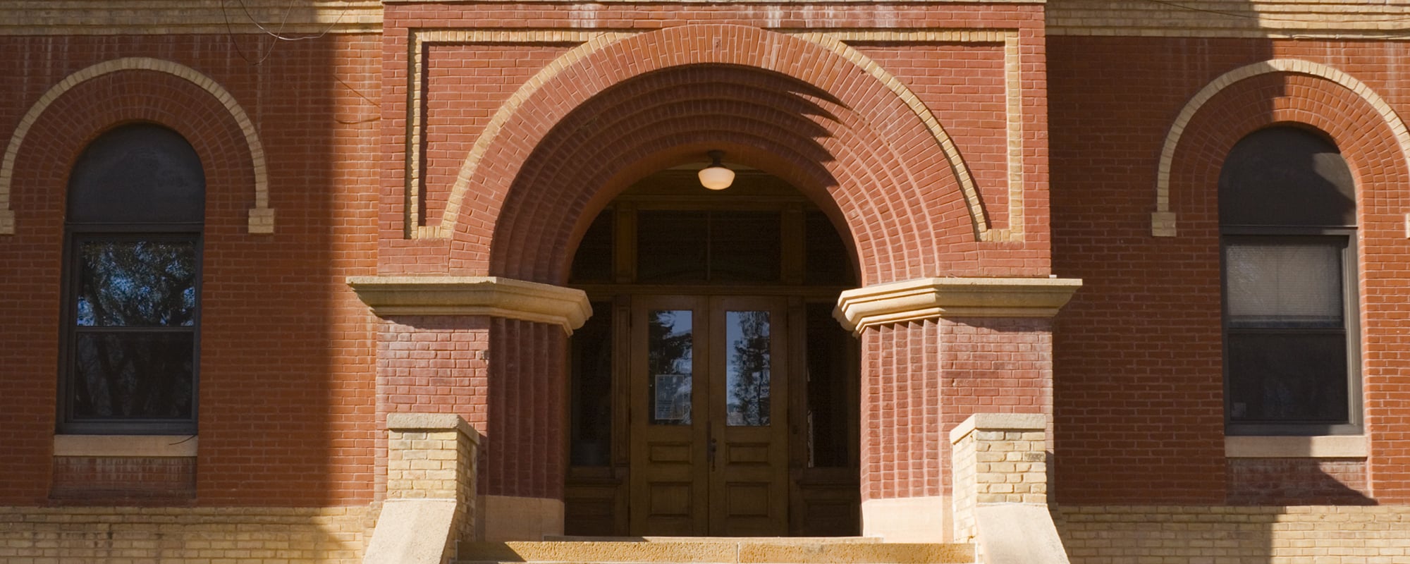 Horizontal straight-on exterior view of an old red brick high school or elementary schoolhouse building facade, centered on the entrance front door and staircase. The traditional architecture suggests a respect for higher education and learning. The brownstone built structure is located in a small Midwestern town in the U.S.A.