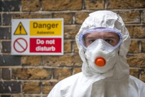 man in full body asbestos personal protective equipment with goggles and mask stands in front of danger sign