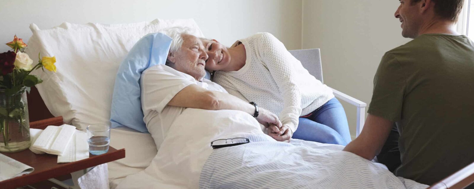 Senior patient lying in hospital bed with family members sitting with him and comforting him.