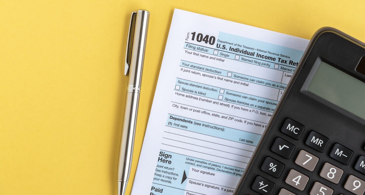 Close up image containing pen, a tax return form, and a calculator on a yellow background.