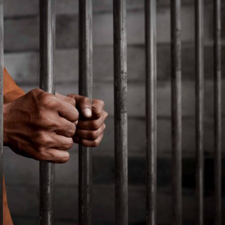 Man in orange prison jumpsuit behind bars in a prison that could have been built with asbestos