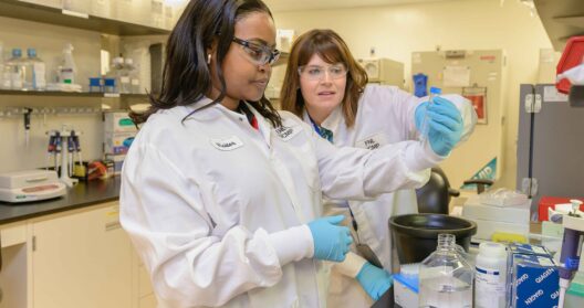 Two female scientists in white lab coats and safety glasses working together in a modern laboratory, examining a test tube sample as part of CAR T-cell therapy research and development